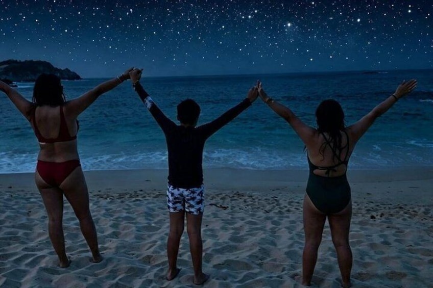 Three people on a beach at night with their arms raised, facing a starry sky. 5