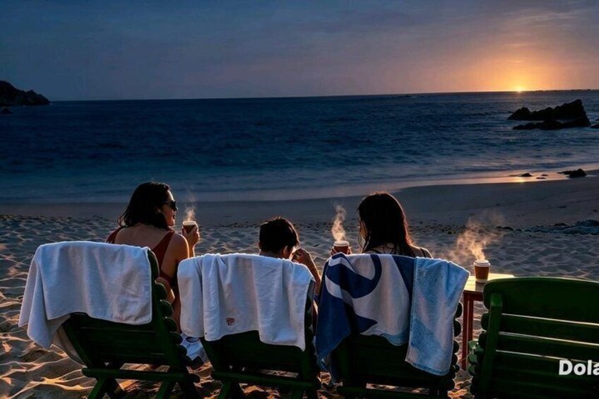 Three people sitting on beach chairs with towels, enjoying a sunset by the ocean. 2