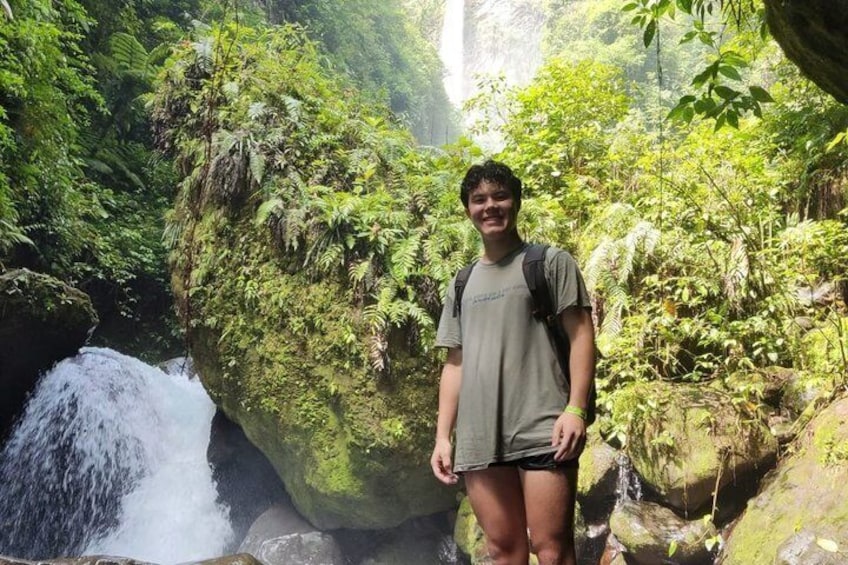 A person standing near a waterfall in a forested area with lush greenery and moss-covered rocks. 5