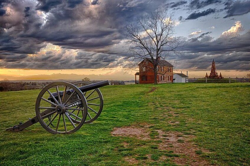 A cannon, a red house, and a church steeple in a grassy field under a cloudy sky. 3