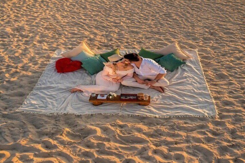 A couple on a beach blanket with pillows and a tray of drinks. 3