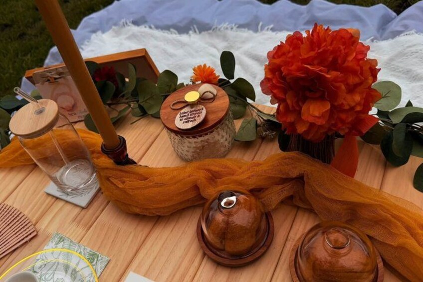 A picnic setup with a wooden table, a glass jar, a wooden spoon, a jar with a lid, a red flower, and a wooden spoon. 1