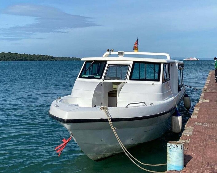 A white boat docked at a pier with a flag on top, a red flag on the bow, and calm water in the background. 2