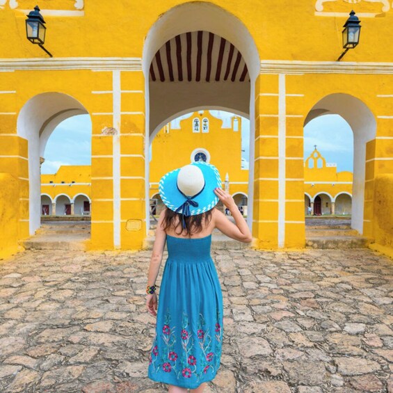 A person in a blue dress and hat walking through a yellow building with arches. 1
