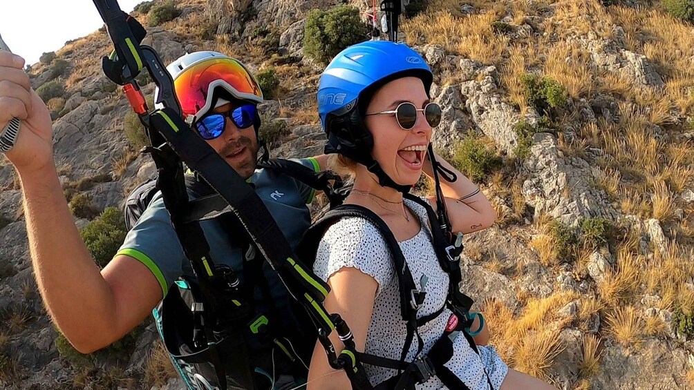Two people with helmets and harnesses, likely engaged in a mountain climbing or hiking activity, with rocky terrain and sparse vegetation in the background. 2