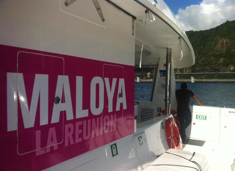 A boat with 'MALOYA LA REUNION' written on its side, a person standing at the stern, and a mountainous landscape in the background. 2
