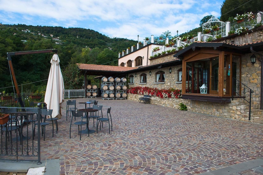 A patio area with outdoor seating, a stone building with wine barrels, and a mountainous backdrop. 1