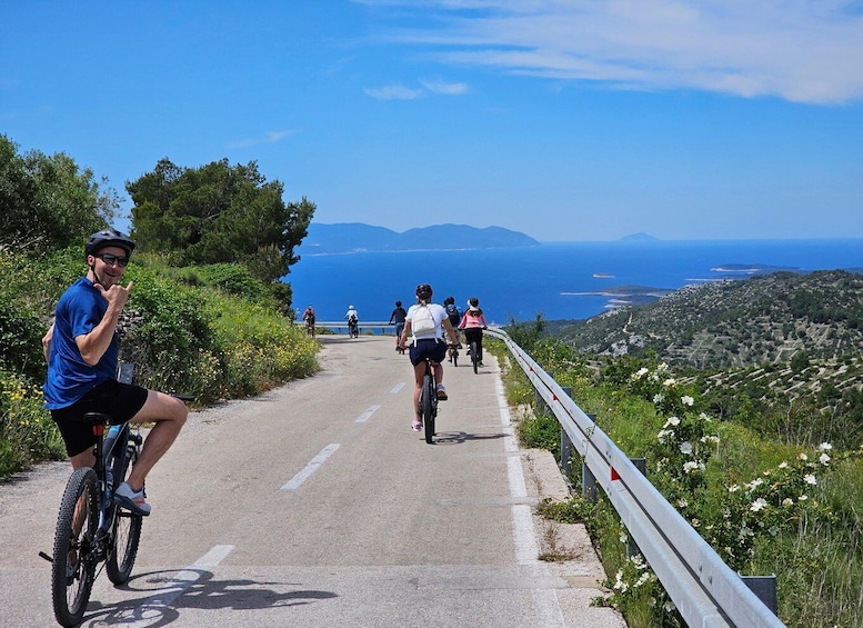Cyclists on a paved road with a guardrail, surrounded by greenery and overlooking a coastal landscape. 3