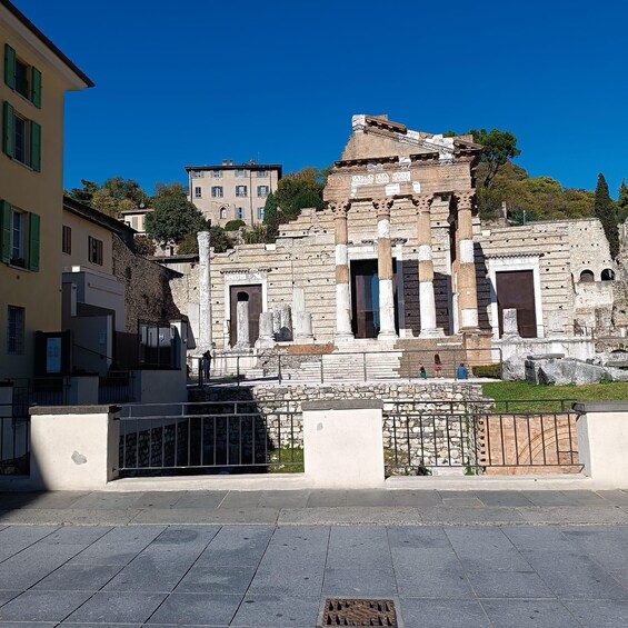 An ancient Roman amphitheater with columns and ruins, surrounded by modern buildings and a clear blue sky. 1