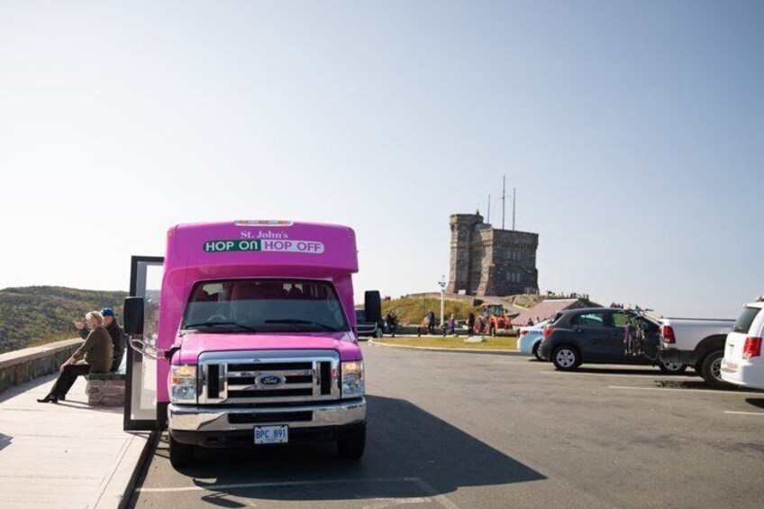 A pink truck with 'St. John's Hop On Hop Off' on the front, parked on a road with a castle-like structure in the background. 1