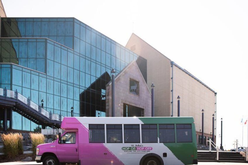 A pink and white bus parked in front of a modern building with glass windows and a staircase. 5