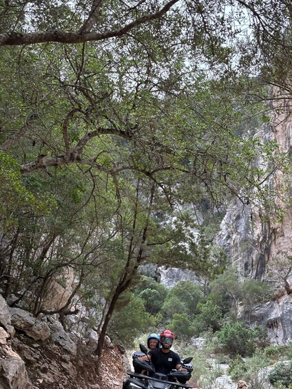 Two people on a quad bike in a forested area with rocky cliffs. 4
