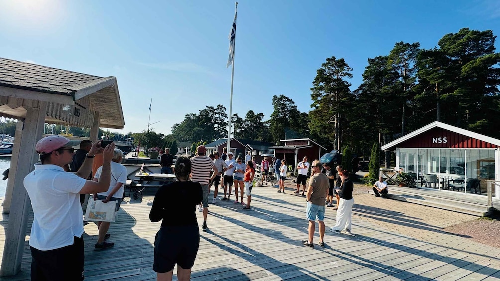 A group of people on a wooden pier, a flagpole with a flag, and a building with 'NSS' on it. 2