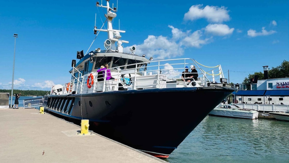 A large black boat docked at a pier with people on board and another boat in the background. 1