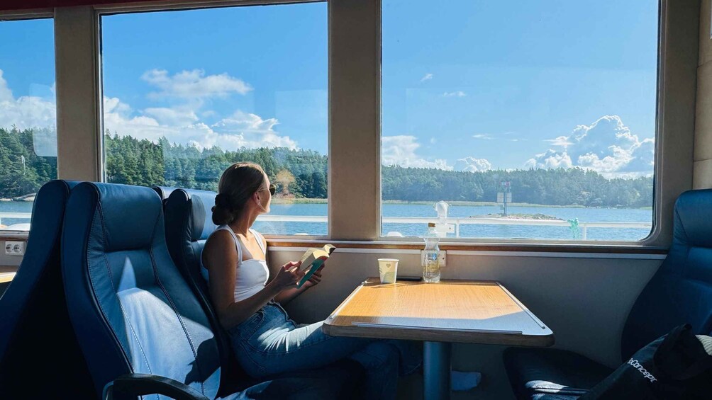 A person seated by a window on a boat, reading a book with a cup beside them. 5