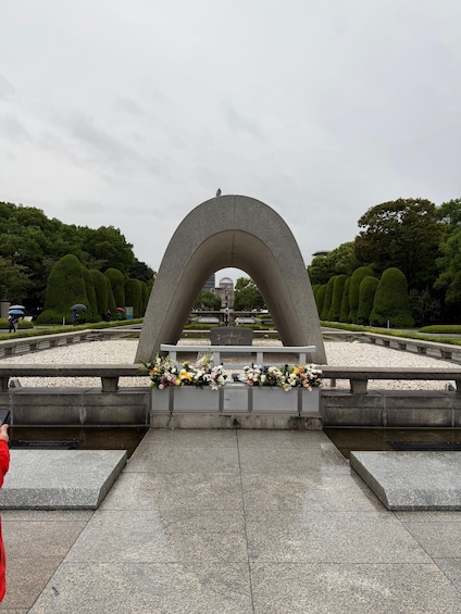 A stone archway with floral arrangements at its base, surrounded by a park with trimmed trees and a distant building. 1