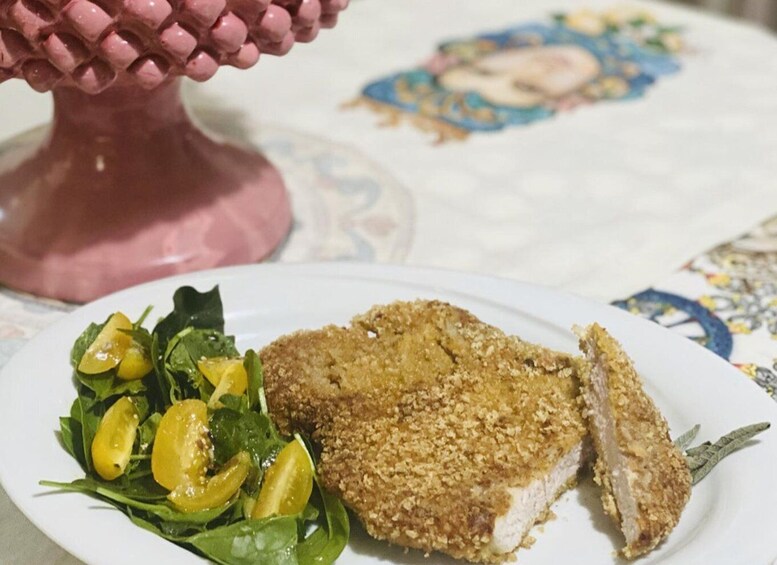 A plate with a breaded and fried cutlet, a side salad with spinach and cherry tomatoes, and a decorative pink object in the background. 4