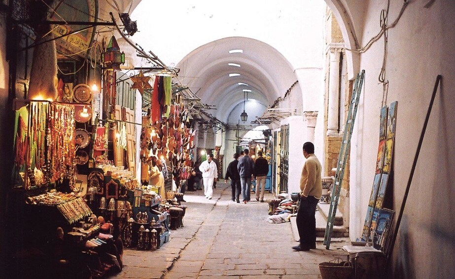 A narrow market street with shops on both sides, people walking, and a man standing with a ladder. 1