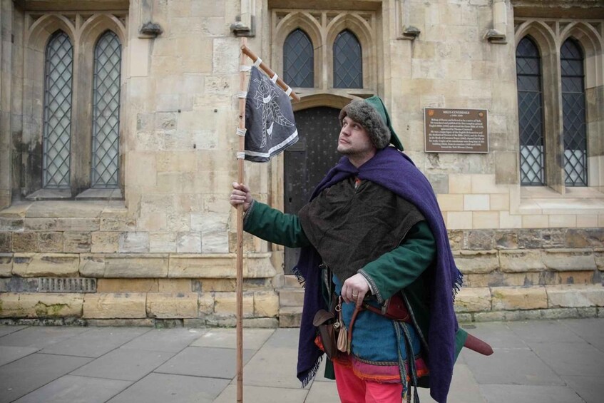 A person in medieval costume holding a flag in front of a stone building with arched windows. 2