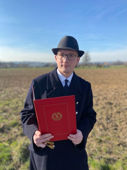 Une personne tenant un livre rouge orné d’un emblème doré, se tenant debout dans un champ. 1
