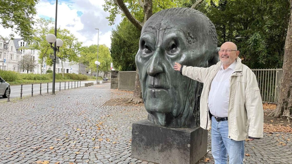 Un homme se tient à côté d’une grande sculpture en bronze représentant un buste, dans un parc aux allées pavées et aux arbres. 3