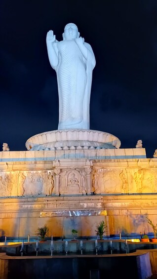 A large white statue with raised hands, set on a pedestal with intricate carvings and a fountain below. 2