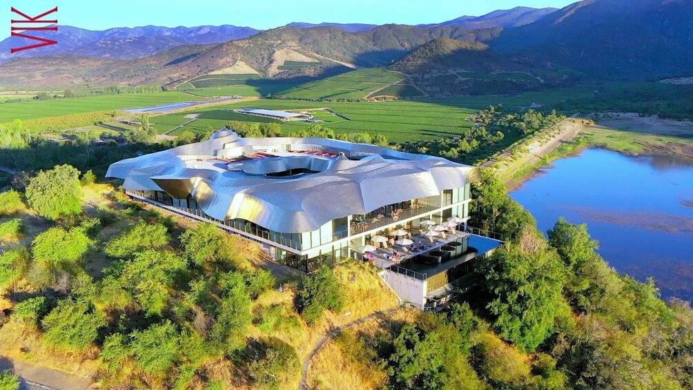 A modern building with a unique roof design surrounded by lush greenery and a lake. 2