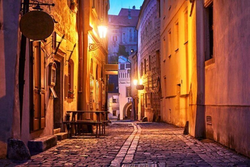 A cobblestone street at dusk with buildings on either side, a bench, and a tower in the distance. 3