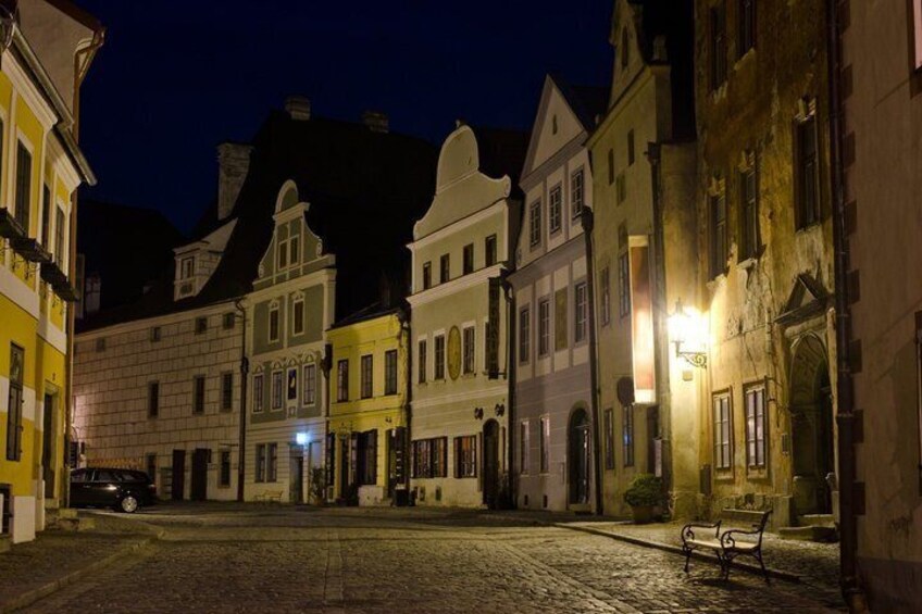 A cobblestone street at night with historic buildings, a bench, and a parked car. 4