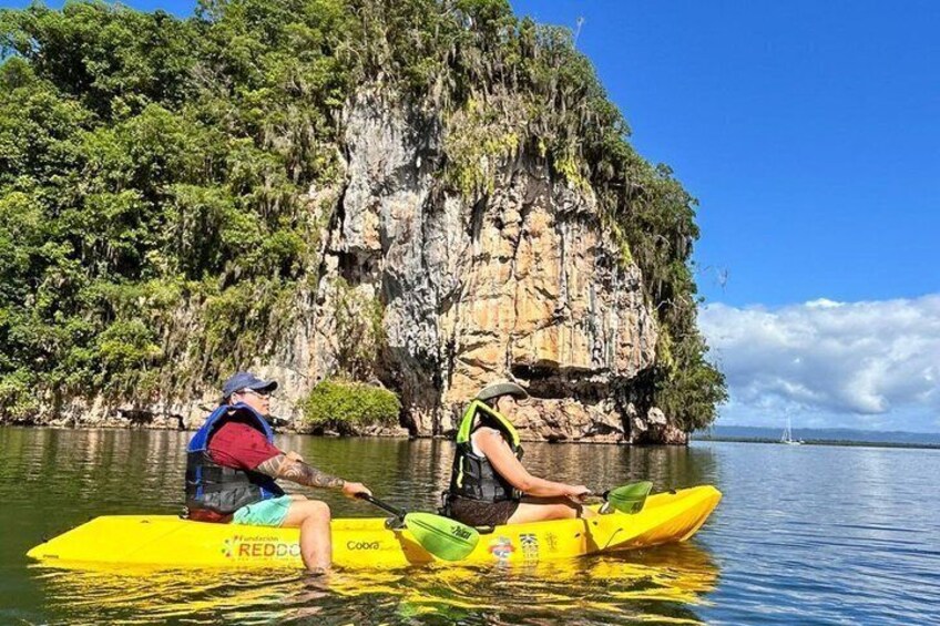 Two people kayaking on a yellow raft in calm water with a rocky cliff and greenery in the background. 3