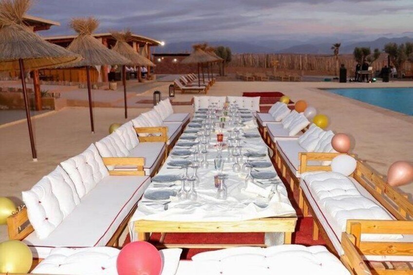 A long dining table set for a meal on a beach with thatched umbrellas and lounge chairs. 3