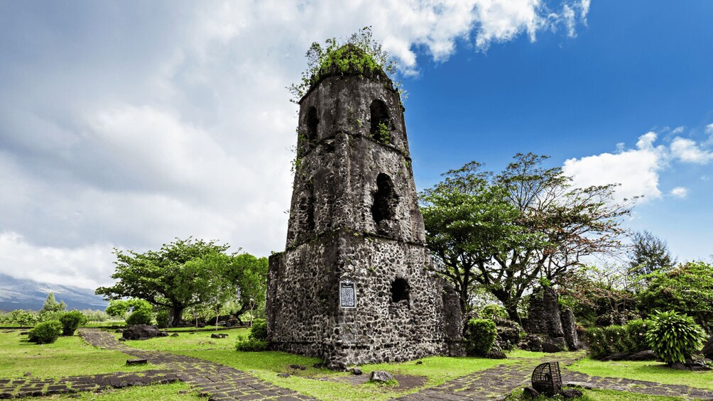 A tall, weathered stone tower with a narrow passageway, surrounded by lush greenery and a clear blue sky. 3