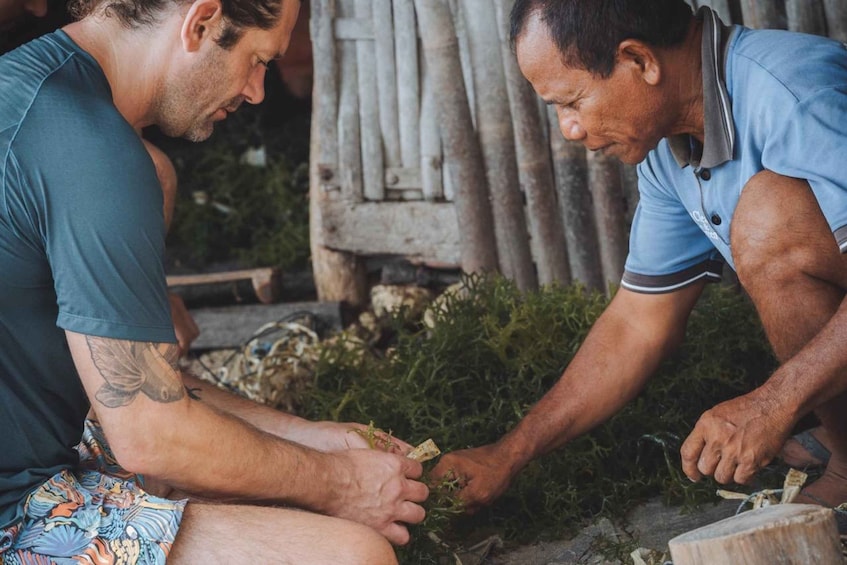 Two men engaged in a manual task, possibly related to agriculture or craft, outdoors with a rustic background including a wooden structure and greenery. 1