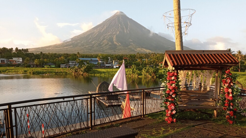 A riverside view with a wooden gazebo decorated with flowers, a mountain in the background, and houses along the riverbank. 1