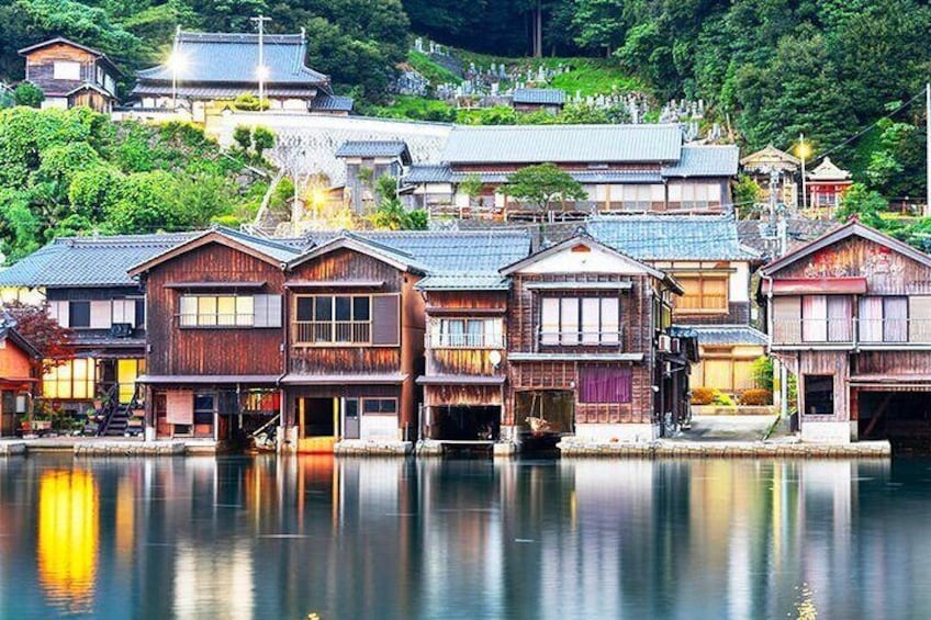 Traditional Japanese wooden houses by a calm river, with a forested hillside in the background. 4