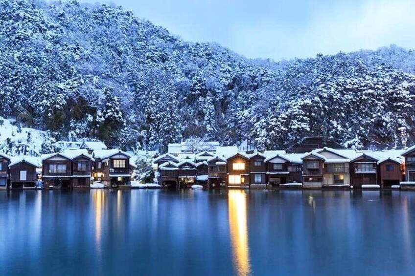 A row of wooden cabins with snow-covered roofs, reflecting on a calm body of water, surrounded by snow-laden trees. 5