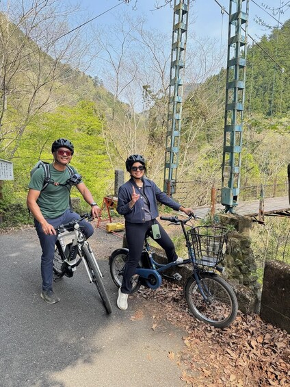 Two people on bicycles, one with a white bike and the other with a blue bike, in a natural setting with trees and a metal structure in the background. 2