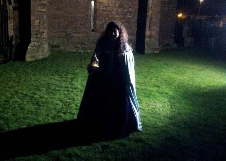 A person in a long dress standing in a grassy area with a brick wall in the background. 1