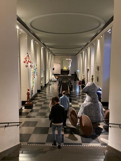 A hallway with a checkered floor, a large sculpture of a figure with a skirt, and a colorful wall art installation. 4