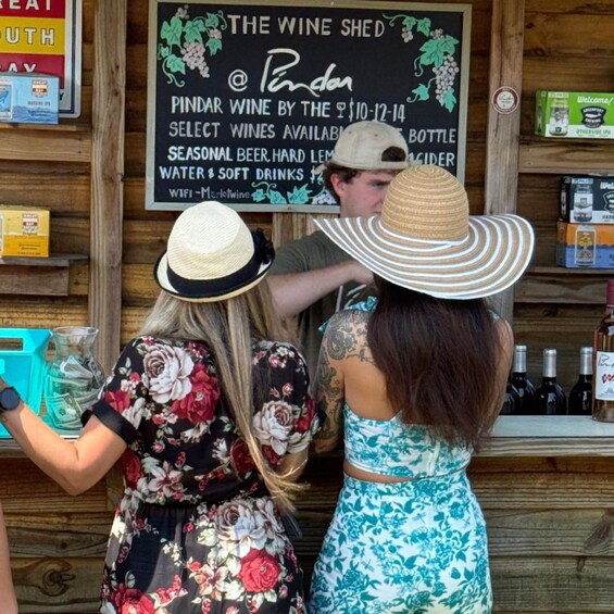 A woman in a floral dress and a straw hat at a wine bar with a sign advertising Pindar wine and other beverages. 3