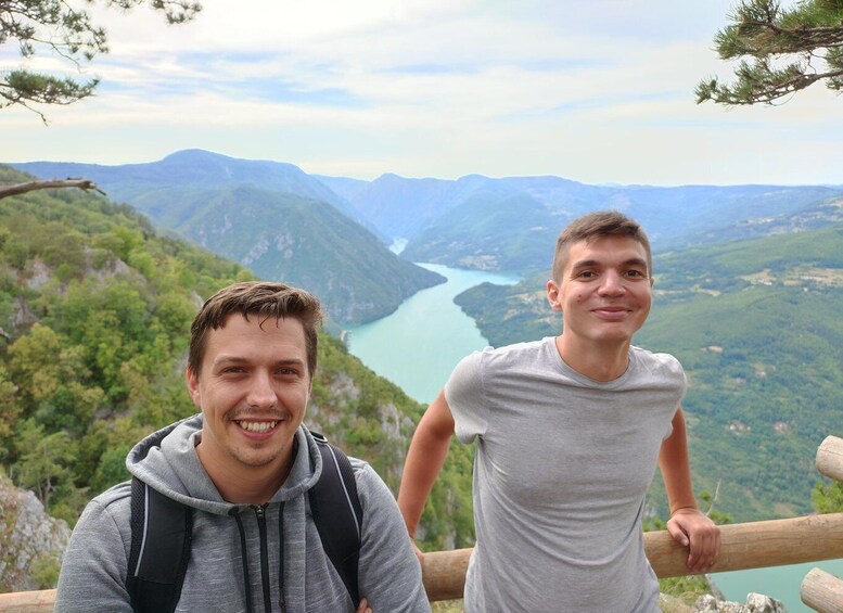 Two people standing on a wooden platform overlooking a river valley with mountains in the background. 3