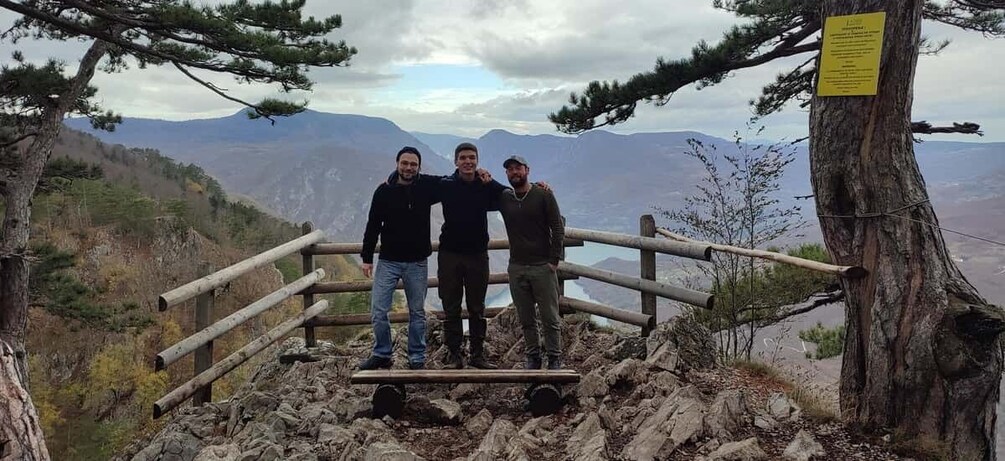 Three people standing on a rocky outcrop with a wooden fence and a sign, overlooking a mountainous landscape. 5