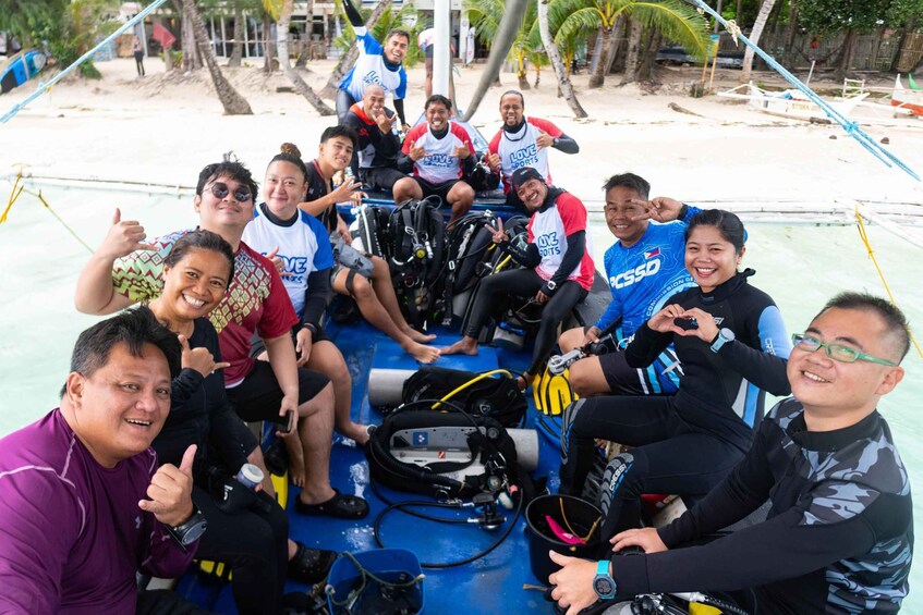 A group of people on a boat with scuba diving equipment, a sandy beach, and palm trees in the background. 3