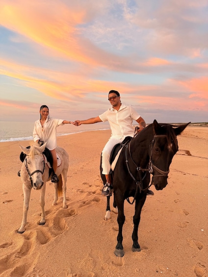 Two people riding horses on a beach with a sunset in the background. 1