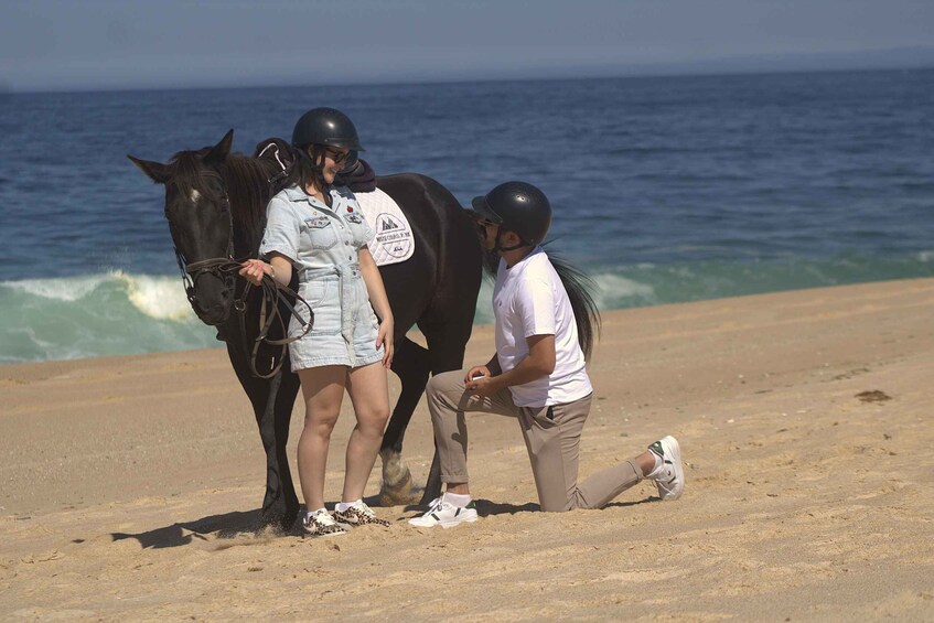 Two people with horses on a beach, one wearing a helmet and the other holding a phone. 5