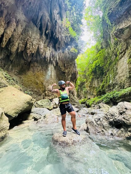 A person standing on a rock in a clear stream with a cave in the background. 2