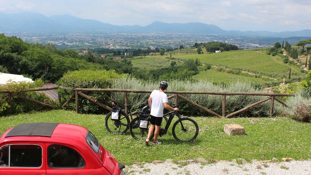A person standing next to a bicycle on a grassy hill with a red car in the foreground and a distant cityscape. 5