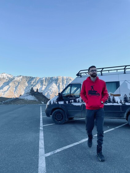 A person in a red hoodie standing in front of a black van with a mountain landscape in the background. 1