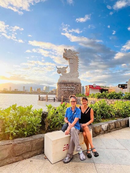 Two people sitting on a bench with a dragon statue in the background and a city skyline across the water. 4