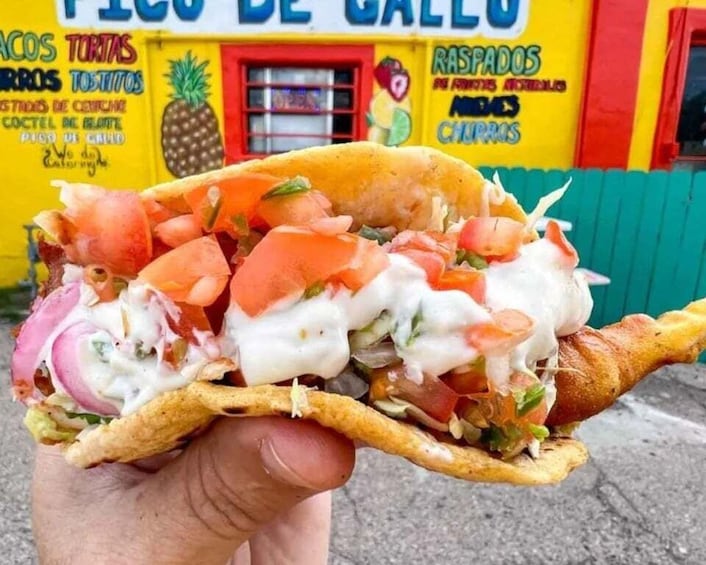 A hand holding a taco with toppings, in front of a colorful food stall. 1